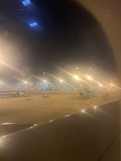 This image captures an airport tarmac at night, likely in Guangzhou, China. The scene is dominated by the bright, star-like glare of runway lights and floodlights illuminating several parked airplanes. The perspective suggests the photo was taken from inside an aircraft, possibly from a window seat.
In the foreground, a dark, curved shape, likely the wing or fuselage of the aircraft the photo is taken from, obscures a portion of the view. Two illuminated "EXIT" signs are visible in the bottom left corner, glowing blue against the dark interior, indicating the location within the aircraft.
Further out, a row of airplanes is parked along the tarmac. Some appear to be passenger jets, with their distinctive shapes and landing gear visible. The terminal building is visible in the distance, with numerous lit windows suggesting activity. Service vehicles, such as a small cart, are also present on the tarmac, contributing to the sense of a busy, functional airport environment even at night.
The lighting creates a high-contrast scene, with bright lights casting long reflections on the wet tarmac. The overall impression is one of a typical airport operational scene during nighttime, with the glow of artificial lights battling the darkness. No people are clearly discernible in the image, but the presence of the terminal and aircraft implies their recent or imminent presence.
