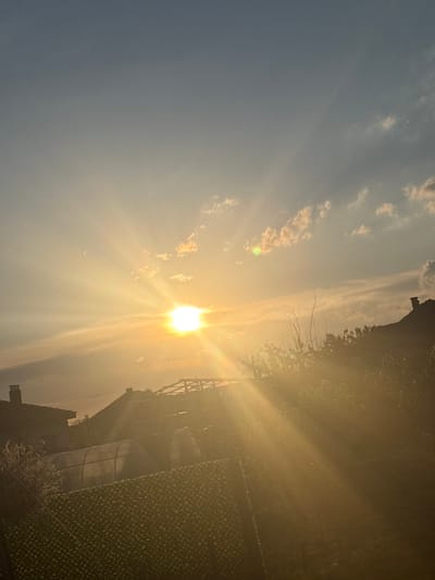 The image depicts a sunlit scene in Kukorevo, Bulgaria. The sun, centrally positioned in the sky, is emitting bright rays, partially obscured by thin clouds. A building or structure with an architectural framework is visible on the right, partially covered in vegetation. A chimney is seen protruding from the building. The sky is a gradient of blue and golden hues due to the sunlight.