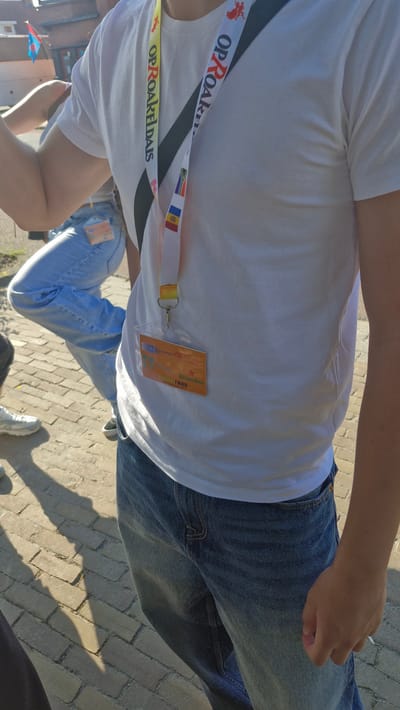 A person wearing a white t-shirt and blue jeans stands outdoors on a paved area. A lanyard with a badge is attached to the t-shirt. The badge is orange and displays text that includes "Heineken" and "VSE Fonds". Another person in blue jeans is visible in the background. There are flags on poles in the upper left corner. The image appears to have been taken on a sunny day.