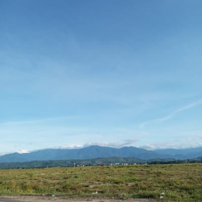 The image presents a view of the landscape in Meunasah Kumbang, Indonesia. The foreground features a field of dry, brownish grass, and a small amount of trash is scattered on the ground. In the midground, there's a series of gently rolling hills and mountains. The sky takes up a significant portion of the image, displaying a clear, light blue hue, with a few wispy clouds scattered across it. The image doesn't show any people.