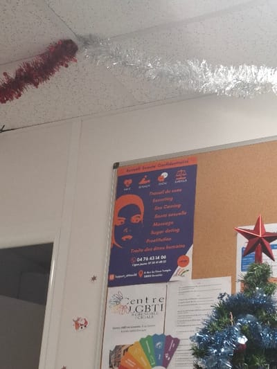The image displays an indoor scene located at 8 rue du Vieux Temple, 38000 Grenoble, France. A white suspended ceiling is visible at the top, adorned with a length of red tinsel and an adjoining length of silver tinsel. Below the ceiling, a white wall is present on the left, decorated with small, silver star stickers and a small Santa Claus sticker. To the right, a cork board is mounted, featuring multiple pinned documents. A prominent blue poster on the cork board is titled "Accueil Écoute Confidentialité" (Reception Listening Confidentiality) and lists services including "Travail du sexe" (Sex work), "Escorting," "Sex Gaming," "Santé sexuelle" (Sexual health), "Massage," "Sugar dating," "Prostitution," and "Traite des êtres humains" (Human trafficking), along with a phone number and the mentioned Grenoble address. Below this poster is another white document identifying "Centre LGBTI #Grenoble / CIGALE," which also includes the 8 rue du Vieux Temple address and displays a fan of rainbow-colored cards. In the lower right corner, a small artificial green Christmas tree, decorated with blue tinsel and topped with a red star, is partially visible. No individuals are present in the scene.