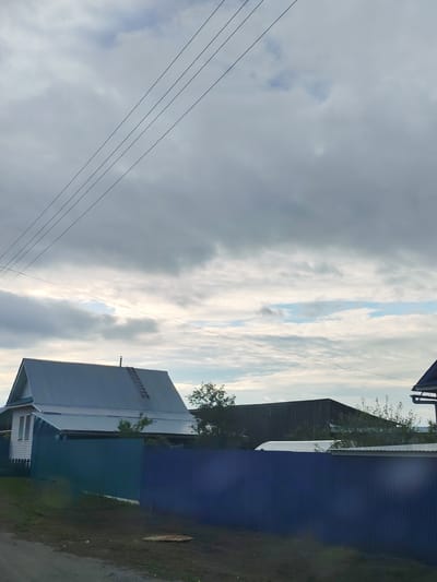 The image depicts a rural or semi-rural scene in Malaya Bod'ya, Russia, under an overcast sky with patches of visible blue. The upper two-thirds of the frame are dominated by gray and white clouds, with a clearer section of blue sky and brighter light appearing towards the mid-right. Multiple thin black power lines traverse the sky diagonally from the upper left, while a thicker, single power line stretches horizontally across the middle of the sky, slightly above the visible structures.
In the mid-ground, a white two-story house with a steeply pitched gray metal roof stands on the left. A wooden ladder is propped against the right slope of its roof, extending from the eave towards the ridge, where a small chimney or vent pipe is visible. An additional, lower gray metal roof extends from the main house. A corrugated metal fence, predominantly dark blue but appearing dark teal on its left section, runs across the mid-ground, separating the foreground from the properties beyond.
To the right of the fence, a dark, presumably wooden, rectangular building with a dark roof is visible behind dense green foliage. A lighter-colored, rounded structure, possibly a covered vehicle or greenhouse, is partially visible behind the fence and in front of the dark building. The edge of another roof with a gutter system is visible in the far upper right corner.
The foreground consists of a dirt or gravel path/road, with green grass and darker vegetation bordering it. A circular metal grate or manhole cover is partially visible on the ground in the lower-right quadrant. No individuals or animals are present in the scene.