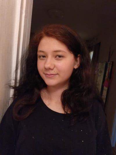 The image is a head and shoulders portrait of a young person with dark, wavy hair that has reddish undertones. The person has light-colored eyes and a mole on their right cheek. They are wearing a dark, long-sleeved shirt with a slightly sparkling pattern. The background is dimly lit, with a textured surface that appears to be a curtain or wall on the left and a darker, indistinct background on the right, possibly containing furniture or a doorway. The lighting suggests an indoor setting, likely at night or in a room with low ambient light. There is no discernible information to indicate the location within the city of Tolbazy, Russia.