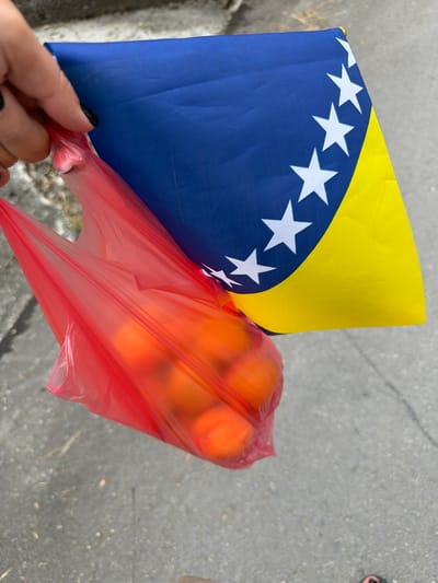 A close-up, outdoor, waist-level shot shows a person's hand holding a red plastic bag filled with what appear to be oranges. The hand, with dark nail polish, is positioned near the top of the bag. Behind the bag is a flag, predominantly blue with a yellow diagonal band along the right side, and white stars scattered across the blue portion. The flag appears to be a small handheld version, possibly a souvenir. The ground beneath is a rough, grey asphalt surface, typical of a street or pathway, with some small pebbles and debris visible. The overall setting suggests a casual, everyday scene, possibly in a public area within the city of Jajce, Bosnia and Herzegovina. The lighting is natural, indicating it is daytime. The focus is on the hand, bag, and flag, with the background slightly blurred.
