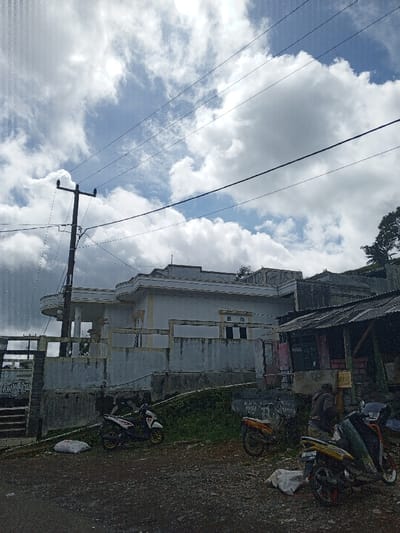 A low-angle shot captures a weathered, multi-tiered building under a cloudy sky. A utility pole and power lines intersect the view. Several motorbikes are parked along the road in front of the building, one of which appears to have someone standing by it. The adjacent structure on the right looks like a simple shop or shelter with a corrugated roof. Overall, the scene evokes a sense of quiet desolation.