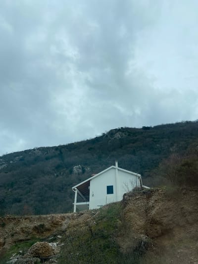 A white building is perched on the edge of a mountain cliff under an overcast sky. The mountain slope is densely covered in vegetation, with bare trees indicating a possible late fall or winter season. A portion of a wall or fence is visible near the building on the cliff edge. The image is taken outdoors in a natural setting. The location is not mentioned in the description.