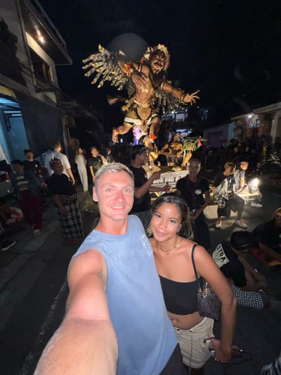 The image displays a nighttime scene on a crowded street in Munggu, Indonesia. In the foreground, a light-skinned male with blonde hair, wearing a blue sleeveless shirt, holds a camera to capture a selfie with a female individual of darker complexion, with dark hair, wearing a black one-shoulder crop top and light cargo shorts, carrying a dark handbag. Both individuals are smiling towards the camera.
Behind the two foreground subjects, a large, ornate effigy, identified as an Ogoh-Ogoh, is being paraded down the street. The primary figure of the effigy is a multi-limbed, winged creature with fangs and claws, adorned with elaborate traditional Balinese gold and brown detailing, positioned in an aggressive posture on a decorative platform. Smaller figures are also visible on the base of the platform.
The street is densely populated with individuals, primarily men, many of whom are wearing traditional black and white checkered sarongs (kain poleng). These individuals are seen carrying the effigy's platform or moving around it. Buildings with multiple stories line both sides of the street, and the scene is illuminated by artificial light sources, casting shadows and highlighting the effigy and foreground subjects. The atmosphere is that of a public procession or cultural event.