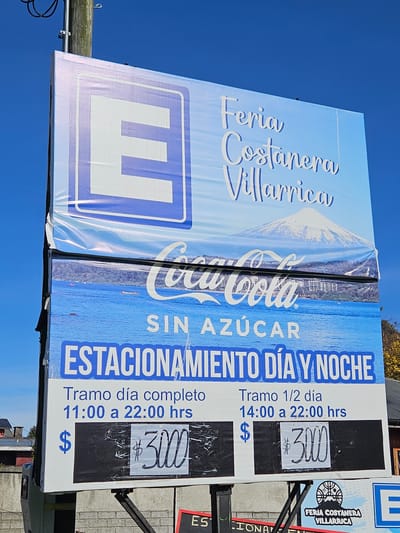 The image shows a sign in Villarrica, Chile, advertising the "Feria Costanera Villarrica" and Coca-Cola Sin Azúcar, the sign also advertises parking, “Estacionamiento Dia Y Noche”, for 3,000 Chilean pesos. "Tramo Dia Completo", “Full Day” from 11:00 to 22:00 hrs, and 3,000 pesos "Tramo 1/2 Dia", “Half Day” from 14:00 to 22:00. The signs are stacked, with the "Feria Costanera Villarrica" and an "E" parking symbol at the top, followed by the Coca-Cola advertisement and parking information. The background is a clear blue sky.
