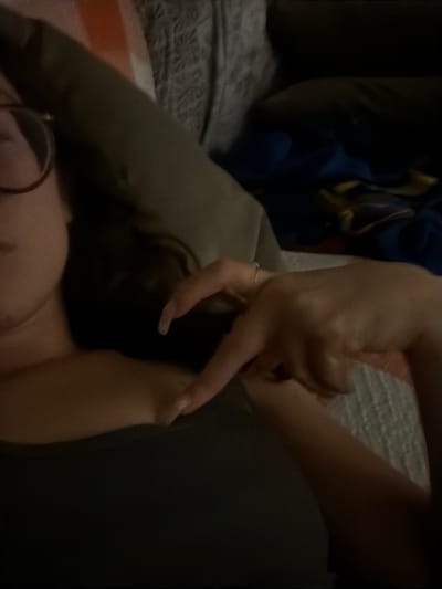The image displays a close-up, low-light, indoor scene featuring a person reclining. A female individual, partially visible from the upper chest, appears on the left side of the frame, wearing a dark-colored top. She has dark hair and wears circular-framed glasses. Her right hand is raised and positioned centrally, with the index finger extended and pointing towards the left, while the thumb is bent and the remaining fingers are curled inward. A slender ring is visible on her right ring finger, and her fingernails are long and appear to be painted a pale color. The background consists of light-colored, possibly striped, bedding or cushions on the upper left, transitioning to dark, blurred fabrics with hints of blue and yellow on the right. The scene is softly lit, contributing to a slight blur and underexposed appearance.