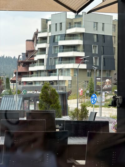 The image shows a modern apartment building with a unique architectural design. The building features a mix of dark gray and light gray facade panels, with white balconies and large windows. To the left of the building, there is a brown wooden structure, possibly a cafe or restaurant, with an awning. In the foreground, dark reflective surfaces, likely glass or polished metal, obscure parts of the scene, creating a layered effect.
Behind the apartment building, there are signs and street furniture. One billboard advertises "Telekom" with an image of two people wearing helmets. A blue circular sign with white arrows indicates a roundabout or a specific traffic flow. Another sign has red lettering and a stylized graphic. Further back, there is a building with a sign that reads "INVICTA Rez." and "PRODAJA" with a price of "291". Green trees and foliage are visible, and in the distance, a densely forested hill is seen under a cloudy sky. The overall impression is an urban or semi-urban setting, likely in the daytime, with overcast weather. The image is captured from a slightly elevated, angled perspective, with the scene appearing rotated.