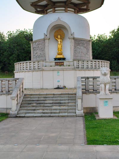 The image shows the Peace Pagoda in Willen Lake, Milton Keynes, United Kingdom. The pagoda is a white, multi-tiered structure with a distinctive roof. In the central niche of the pagoda, a golden statue of the Buddha is displayed. Relief carvings depicting scenes from Buddhist mythology are present on the lower tiers of the pagoda. In front of the pagoda, there are stone steps leading up to a circular walkway bordered by a stone balustrade. To the right of the steps, a stone statue of a lion stands on a pedestal. The surrounding area is a grassy lawn with trees in the background. The sky is clear and pale blue, suggesting it is daytime. The overall scene is serene and spiritual, reflecting the purpose of the Peace Pagoda as a monument to peace.