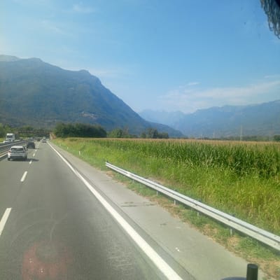 The image depicts a highway scene with mountains and a cornfield in the background. The perspective is from a vehicle moving along the highway, as indicated by the road markings and the slight motion blur. Several cars are visible on the highway, including a white SUV in the foreground. A truck is seen in the distance on the left. The highway has multiple lanes, separated by white lines. A metal guardrail runs along the right side of the road, adjacent to a vibrant green cornfield. In the background, lush green mountains rise under a clear blue sky. The image appears to be taken during daylight, with the sun casting shadows. The specific location in Calea, Italy, is not explicitly identifiable from the visual cues alone, as the scene is characteristic of many highways in mountainous regions of Italy.