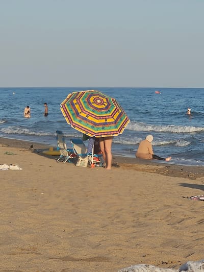 A bright, colorful umbrella is open on a sandy beach, providing shade for people lounging in chairs. The sea is a vibrant blue with gentle waves lapping at the shore. Several individuals are visible in the water, either swimming or wading. One person is lying on the sand, partially covered by a beige garment and hat, facing the sea. The scene suggests a sunny day at the beach, likely in the afternoon given the lighting. The sand shows ripples from the receding tide, and there are a few scattered items like beach chairs and bags. The overall mood is relaxed and leisurely, indicative of a summer vacation. The location is identified as Castelldefels, Spain.