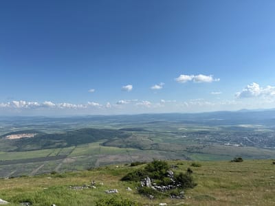 The image presents a wide-angle panoramic view from a elevated position, likely a hilltop or mountain, overlooking a vast landscape under a clear blue sky. Approximately two-thirds of the upper frame is occupied by a bright blue sky with scattered white cumulus clouds of varying sizes; a denser cluster is visible on the far right horizon, while smaller, more isolated clouds dot the central sky.
The immediate foreground, comprising the bottom 15-20% of the image, consists of a gently sloped grassy terrain. The grass is a mix of vibrant green and drier, yellowish-green patches. Numerous grey-white rocks and small, sparse green bushes are distributed across this area, with some rock formations appearing as loose piles of stone in the lower center and left.
Beyond the immediate foreground, the midground and background reveal an extensive rural and semi-urban landscape. In the mid-left, a large, light-colored open-pit quarry with distinct terracing is visible, surrounded by dark green forested areas. Adjacent to this, and stretching across the center of the image, are numerous agricultural fields, predominantly green, some exhibiting rectangular patterns and others irregular shapes, suggesting various crops or stages of cultivation. Darker green forest patches are interspersed among these fields and cover some low hills.
Towards the mid-right and extending to the right edge, a compact town or city is clearly visible, comprising a dense cluster of buildings with visible road networks. This settlement is identified as Dragoman, Bulgaria. Further into the background, successive layers of hills and mountains recede into the hazy distance, appearing bluer due to atmospheric perspective. No individuals, animals, or active interactions are discernible within the frame. The scene is illuminated by bright, natural sunlight, indicating fair weather conditions.