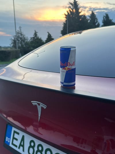 The image captures a static scene in Momkovo, Bulgaria, at twilight, likely sunset or sunrise. A red Tesla vehicle is prominently featured from a rear-three-quarter perspective. A standard 250ml Red Bull energy drink can, silver and blue with visible Bulgarian text "Освежете студена." and "Съживява тялото и ума.", stands upright on the car's rear spoiler. The Tesla's emblem is visible on the trunk, below which a Bulgarian license plate (BG EA 8802 EA) is partially legible. The car's surface, including the trunk and rear window, displays small water droplets. The background consists of a vibrant sky transitioning from orange and yellow at the horizon to blue overhead, with silhouettes of several dark coniferous trees visible against the lighter sky. A slender, vertical pole or antenna is present on the left side of the frame. No individuals are depicted in the scene.