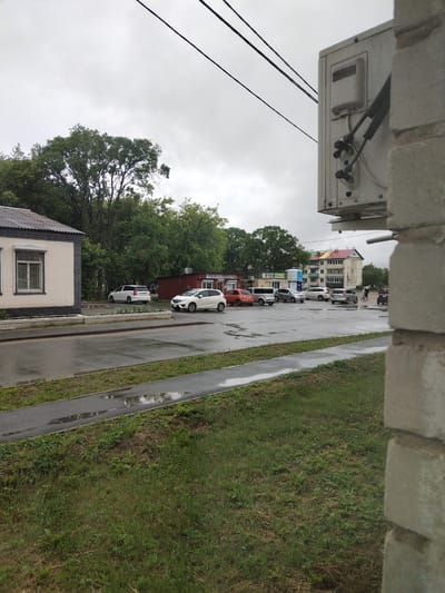 The image depicts a street scene in Pokrovka, Russia, on an overcast day. A wet asphalt road occupies the foreground, with a strip of grass and a paved walkway on the left side. Several cars are parked in a lot across the street, including a white SUV, a silver hatchback, and a small orange car. In the background, a multi-story building with a brightly colored facade stands against a backdrop of trees. An air conditioning unit is mounted on the right side of the frame, partially obscuring a brick wall. The overall impression is one of a quiet, possibly residential, urban environment.