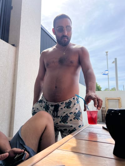 The image shows two men sitting at a wooden table. The man standing in the foreground is wearing a pair of white swim trunks with a blue and green turtle pattern. He is shirtless and has a beard and sunglasses. He is holding a glass of red liquid with a blue straw in his right hand. He is looking down at the glass, which is resting on the table. The other man is sitting at the table, only his legs are visible. The image is taken outside on a patio. The table is located in the city of Kfar Yona, Israel (latitude: 32.317, longitude: 34.947). An Israeli flag can be seen in the background. The image is slightly out of focus. The lighting is bright, likely midday sun. The image is shot from a low angle, looking up at the men. The man in the foreground appears to be taking a drink from the glass. There are various unidentified objects in the background, including a black object that looks like a phone or a camera lens, potted plants, and a bamboo frame. There is also a white wall and a metal fence in the background.