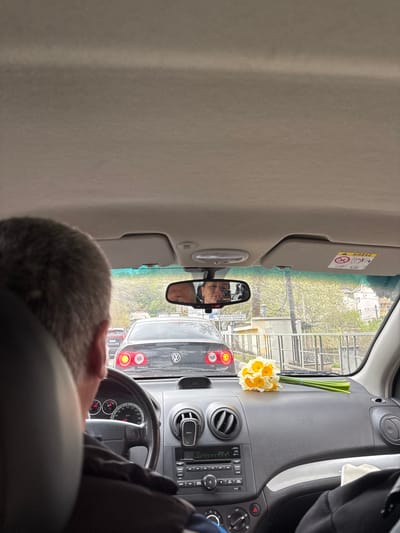 The image captures a vertical perspective from inside a car, looking forward through the windshield. In the bottom right foreground, the back of a person's head with short, greyish hair is visible, suggesting they are in the driver's or front passenger's seat.

The car's dashboard and steering wheel occupy the right side of the frame, showcasing air vents, a radio control panel, and a hazard light button. A cheerful bouquet of yellow and white daffodils, bound by a light blue elastic band, rests prominently on the dashboard near the windshield on the far right.

The rearview mirror, positioned in the upper center, reflects the face of an older man with short hair, likely the same individual seen from behind. Through the windshield, an outdoor scene unfolds. The car appears to be stationary, possibly in traffic, as the rear of a dark-colored car is directly ahead. Text on its trunk identifies it as a "PASSAT," and its red taillights are visible. Beyond this vehicle, trees with fresh green foliage hint at spring or early summer, alongside glimpses of buildings and fences.

An "AIRBAG" warning label is clearly visible on the sun visor in the upper left. Further contributing to location cues, Russian text is partially discernible on a sign reflected in the rearview mirror, including fragments like "...АДНЫЙ РАСКОМЫС..." and "...p. РАДУМЫЙ РАБОМЪ". The natural lighting suggests it is daytime. The scene conveys a tranquil moment within an urban or suburban setting in Bolshoy Sochi, Russia.