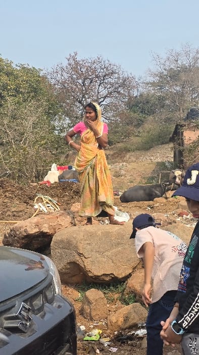 A woman wearing a pink top and patterned yellow sari stands in a rocky, trash-strewn area near bare trees. A dark buffalo grazes behind her. Two children wearing hats and casual clothing stand to the right, near a large rock and a dark SUV. The scene depicts a rural landscape with visible environmental concerns.