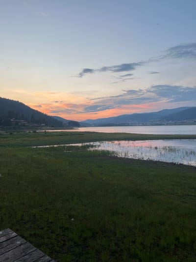 The image depicts a serene lakeside scene at dusk in Sarnitsa, Bulgaria. The sky is a gradient of soft blues and pinks, with wispy clouds scattered across the horizon. A distant town with buildings is visible nestled against rolling hills on the far side of a calm lake. The foreground is dominated by lush green grass that slopes gently towards the water. Reeds and aquatic plants are present in the shallower parts of the lake. A wooden dock or pier is partially visible in the lower left corner of the frame, suggesting a recreational area. The overall atmosphere is peaceful and tranquil, with the soft light of sunset casting reflections on the water.