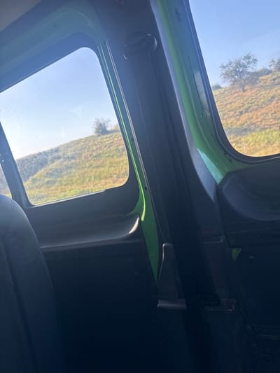 The image is taken from inside a vehicle, looking out of a window towards a grassy, hilly landscape. The vehicle's interior is visible, including a dark-colored seat, the green frame of the window, and part of the door panel with a seatbelt. The exterior shows a sunny sky, rolling hills covered in dry grass and some scattered trees. The location is identified as Dobrotitsa, Bulgaria, suggesting a rural or semi-rural setting. There are no people visible in the image. The overall impression is one of travel through a dry, possibly arid or seasonally dry, rural environment.