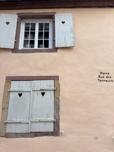 The image shows the facade of a building in Colmar, France, with two windows. The upper window has white shutters with heart cutouts, and the lower window also has white shutters with heart cutouts. A street sign reading "Petite Rue des Tanneurs" is visible on the wall to the right of the windows. A small plaque is also attached to the wall below the street sign. The building has a pale pink stucco exterior and a dark wooden roofline.