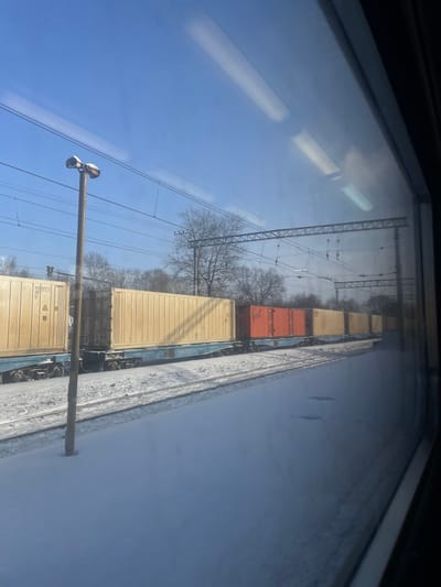 The image displays a snowy railway scene in Elektrougli, Russia, observed through a train window. A long freight train occupies a parallel track on the left, composed of multiple blue flatbed railcars. These railcars carry various intermodal shipping containers; the majority are light yellow or tan, with one prominent orange container situated in the middle of the visible train section. Markings "COH 957303" are discernable on a yellow container near the front, and the number "51097583" is visible on a blue railcar. The ground, including the tracks and an adjacent snow-covered platform or open area, is blanketed in white snow. A tall, light-colored lamp post with two top-mounted fixtures stands in the left-mid foreground. Numerous overhead electrical lines and metal gantry structures span the tracks against a clear blue sky. Deciduous trees, bare of leaves, line the distant background. Reflections of internal light fixtures are visible on the window pane. No individuals are present, and no actions are explicitly depicted.