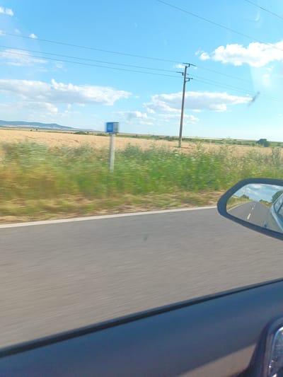 The image is a photograph taken from a moving vehicle on a sunny day. It depicts a rural landscape with a road, a field of dry grass or crops, and a utility pole. In the foreground, the side of the vehicle and a portion of the dashboard are visible. A rearview mirror reflects the road ahead, showing a white dashed line dividing the lanes. The sky is blue with scattered white clouds. The overall scene suggests a journey through the countryside. There is no indication of the city of Zimnitsa, Bulgaria.