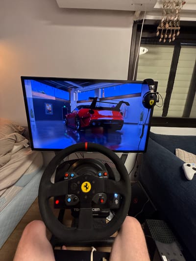 An upside-down interior shot features a gaming setup. A large screen displays a red car in a virtual race. A Ferrari-branded steering wheel is prominently positioned. A headset also bearing the Ferrari logo hangs from the top of the screen. An Xbox console, and a Dell laptop are visible behind the steering wheel. An Xbox controller lies on a couch in the background. Part of a person's legs can also be seen.