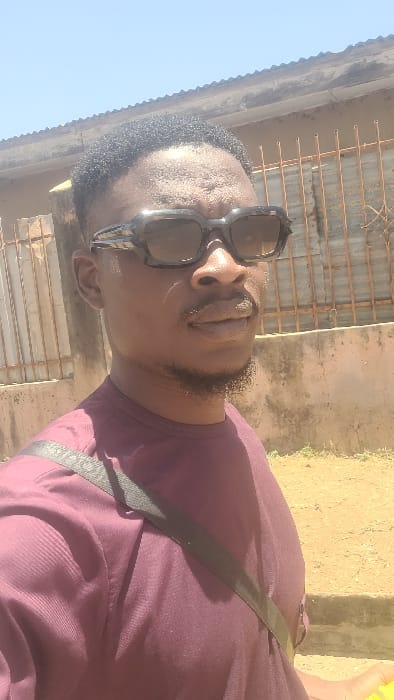 A male subject is depicted in a close-up, waist-up portrait taken outdoors in Badiko, Nigeria. The subject, positioned slightly off-center to the left, has dark skin, short dark curly hair, and a short beard and mustache. He wears dark, rectangular-framed sunglasses that obscure his eyes. His attire consists of a maroon long-sleeved shirt. A black strap, consistent with a shoulder bag, crosses his chest diagonally from his left shoulder towards his right hip. The subject is looking towards the right side of the frame, past the camera. The background reveals a sunlit environment: a light-colored, dusty ground surface in the foreground, a faded beige concrete wall in the mid-ground, and a section of a rusty metal grate or fence above it. A portion of a building with what appears to be a light-colored roofline or eaves is visible in the upper background against a clear, light blue sky. Strong daylight is indicated by visible shadows on the subject's face and clothing.