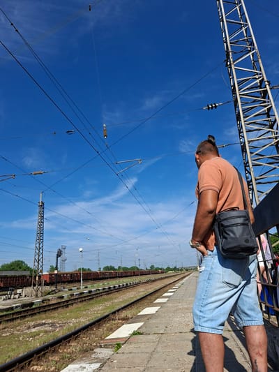 The photograph is taken from a low angle, looking upwards and slightly along a railway platform. In the foreground, a person stands on the platform, wearing a peach-colored t-shirt and light blue denim shorts. They have a dark-colored shoulder bag slung across their body. The person is looking downwards, possibly towards the railway tracks. The platform is made of concrete slabs with white and red markings along the edge.
Parallel to the platform are railway tracks running into the distance. There are train cars or carriages visible on the tracks. Between the tracks and the platform, there is an area of grass and small plants. Overhead, there is a complex network of wires and metal structures, typical of electrified railway lines. The background is a clear blue sky with wispy clouds.
The location is likely Levski, Bulgaria, given the geographical hint. The scene depicts a typical railway platform with elements of the infrastructure required for electric trains.