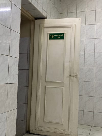The image shows a partially open white door against a tiled wall. The door has decorative paneling and a metal handle. On the door, a green sign with white text reads "19 ВАРОЧНЫЙ ЦЕХ". This translates to "19 Boiling Workshop". The door opens into a dark, recessed area which appears to be a pit or a low-lying room. The surrounding area is also tiled with white and grey tiles. The scene appears to be indoors, possibly in an industrial or utility setting, given the sign. There are no people visible in the image. The lighting suggests it could be daytime, but the lack of windows or external views makes it difficult to determine the time of day or weather. The location is specified as Ranchpar, Armenia, which adds context to the potentially industrial nature of the scene.