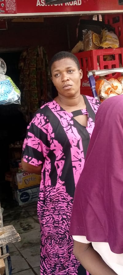 A woman with short hair wearing a pink and black patterned dress stands near a shop entrance. A person in a maroon garment is partially visible on the right. Shelves and goods are visible inside the shop, including bread, drinks, and other packaged items. "ASHTON ROAD" is written on the shop front. A QR code sticker is also affixed to the shop facade. The woman appears to be looking off to the side, with a neutral expression.