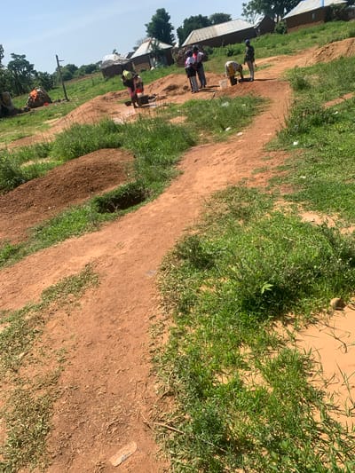 The image depicts an outdoor scene in Buga da Gindi, Nigeria, characterized by a dirt path winding through an area with scattered huts, piles of earth, and vegetation. In the middle ground, several individuals are engaged in activities around a shallow well or water source. One person, wearing a yellow head covering and pink clothing, is bent over, seemingly drawing water. Nearby, three other individuals stand or crouch, with one person in a white top and patterned skirt is also interacting with the water source. Further in the background, a cluster of traditional huts with corrugated metal and thatched roofs are visible amidst trees. The ground is predominantly red earth, with patches of green grass and scrub. To the left, a pile of what appears to be firewood or building materials is stacked near a wooden pole. The overall impression is one of daily life and resource gathering in a rural Nigerian settlement.
