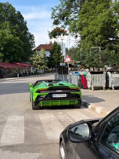 A bright green convertible Lamborghini is parked on a street in Barziya, Bulgaria, facing away from the camera. The car is positioned on a pedestrian crossing, with its rear bumper and taillights prominently displayed. The license plate reads "PB 9709 PE".

To the left of the car, a building with a red-tiled roof and outdoor seating areas is visible, suggesting it may be a cafe or restaurant. Behind this building, trees and a white structure with a red roof can be seen.

To the right of the Lamborghini, several large, grey, industrial-style trash bins are lined up along the sidewalk. A red and white triangular warning sign with a pedestrian symbol is posted on a pole near the bins.

In the foreground, the side mirror and fender of a black car are visible, indicating it is also parked on the street, possibly behind the Lamborghini. The environment appears to be a sunny day, with a blue sky and green trees overhead. The street is paved, and there are white lines marking the pedestrian crossing.