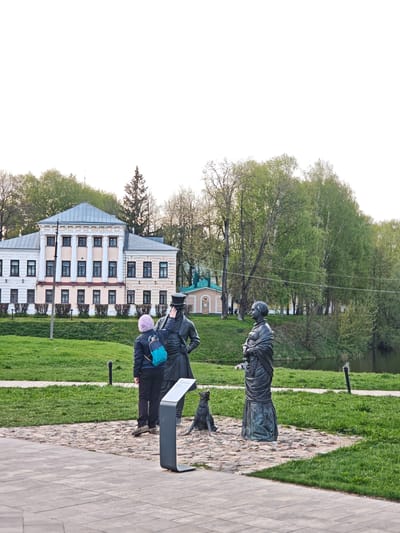 A medium, eye-level shot shows a statue of a couple with a dog in Uglich, Russia. A woman in a blue jacket and pink hat stands next to a statue of a man wearing a top hat. She is reaching up and touching his hat. To the right, a statue of a woman is wearing a long dress. A bronze dog statue sits near the statue of the man. A black metal sign is in front of the man. Behind the statues is a paved stone path leading to a green lawn. A building with a gray roof and white walls is behind the lawn. Green trees surround the building. A small pond is behind the trees on the right. The sky is white and overcast.