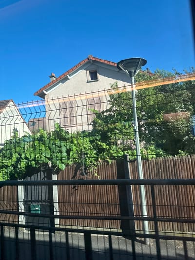 This is a photograph taken from a moving vehicle, likely a train, looking out a window. The image shows a residential area with houses, a fence, lush green foliage, and a street lamp. The houses have pitched roofs with terracotta tiles and light-colored walls. One house has shutters on its windows. The fence is made of vertical wooden planks. In the foreground, a wire fence casts shadows on the ground, indicating the presence of sunlight. A green mailbox is visible behind the fence. The sky is clear and blue. The scene is set in Sèvres, France.