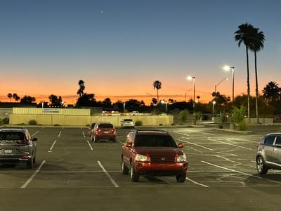 The image depicts an outdoor parking lot at dusk in Phoenix, Arizona, United States. The sky exhibits a gradient from deep blue at the top to a prominent orange and peach hue near the horizon, with a single bright celestial object visible in the upper left quadrant. Multiple artificial streetlights illuminate the asphalt parking surface, which shows white painted lines delineating parking spaces and some visible surface cracks.
Six vehicles are present:
1. A dark red SUV, possibly a Toyota Highlander, is positioned centrally in the foreground, facing towards the viewer and partially occupying a parking space.
2. The rear of a dark gray SUV, likely a Honda CR-V with license plate "9FABCHA," is visible on the far left, parked perpendicular to the camera.
3. The rear of a silver SUV, possibly a Kia Sportage, is visible on the far right, parked perpendicular to the camera.
4. The rear of a dark red sedan is parked in the mid-ground, left-center, facing away.
5. The rear of another dark red sedan is visible behind the first sedan, partially obscured.
6. The rear of a light-colored sedan is parked further back in the mid-ground, center-right.
Numerous palm trees and other silhouetted trees are visible across the horizon, characteristic of a desert environment. In the background on the left, a long, light-colored modular structure or container system is present, bearing a sign with text "CMG SOUTHWEST" and "800.685.9194" partially visible. No individuals are present, and no active interactions are depicted.