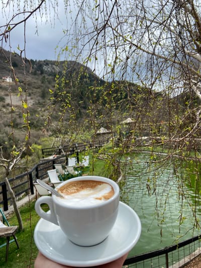 The image shows a white cappuccino cup on a saucer, held in a hand. The cup contains coffee with foamed milk. A spoon is visible in the cup. The background includes a body of water, trees with budding leaves, and a mountain in the distance. There are chairs visible near the water's edge, along with a black metal fence. The photograph appears to be taken at an outdoor cafe. While the location is described as Krapina, Montenegro, there's no information within the image to verify that location, or the presence of a city there.