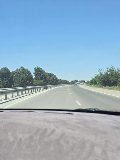 The image shows a view from inside a vehicle traveling on a highway. The highway is paved with asphalt and has white lane markings. A metal guardrail runs along the left side of the road. On the left side of the highway, there is a line of trees. On the right side of the highway, there are also trees and some vegetation. The sky is clear and blue. In the distance, there are a few vehicles on the road, including a white truck and a white car. The foreground of the image shows the dashboard of the vehicle, which is gray and textured. This scene is consistent with a highway in the city of Rabatiohun, Uzbekistan, under a clear sky during the day.