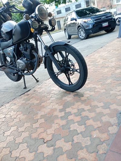 A black Bera motorcycle is parked on a sidewalk paved with interlocking gray and beige blocks. A black Ford Explorer is parked on the street behind the motorcycle. The location is Barquisimeto, Venezuela.