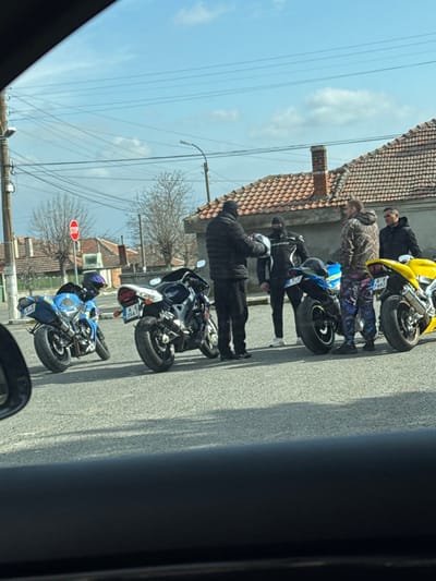 In Vinarsko, Bulgaria, during daytime with a partially cloudy sky, a group of five men are gathered on an asphalt road next to four sport motorcycles. The scene is viewed from inside a vehicle.

On the left, a blue sport motorcycle (Suzuki GSXR, license plate A A 8900) is parked with a helmet resting on its seat. To its right, a dark blue and black sport motorcycle (Honda CBR, license plate A K 3561) is parked. Further right, a blue and white sport motorcycle (Suzuki GSXR, partial license plate B 173) is parked, partially obscured by individuals. On the far right, a yellow sport motorcycle (Suzuki GSXR, license plate A K 7482) is parked.

Among the men, one individual on the far left wears a black puffer jacket, dark pants, and a black balaclava, holding a white motorcycle helmet. Adjacent to him, another man wears black motorcycle racing gear with white accents and a black balaclava. To their right, one man wears a camouflage-patterned hoodie and brightly patterned pants. Two additional men are visible further right, one in a dark jacket and another in a black jacket, both with uncovered faces. The men appear to be conversing or preparing.

The background features a utility pole with multiple overhead wires, bare deciduous trees, and several single-story houses with orange tiled roofs. A red octagonal STOP sign is visible on a pole to the left. A brick chimney is prominent on one of the house roofs.