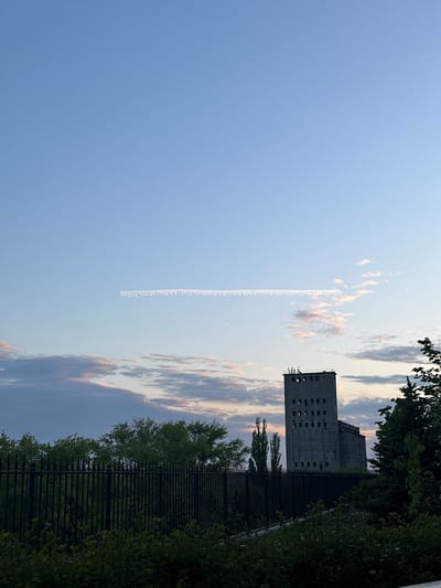 The image shows a blue sky with a contrail extending vertically. There are scattered clouds with a hint of pink and orange. A concrete building with multiple window openings is located on the right side. The building is partially obscured by green foliage and a black metal fence. The location is the city of Kuleshovka, Russia.
