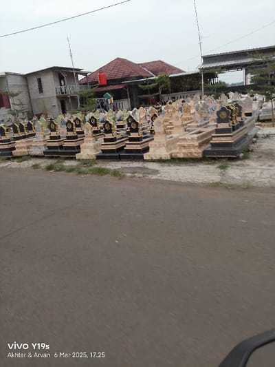 The image shows a street in Kecamatan Gabuswetan, Indonesia (latitude: -6.43, longitude: 108.012) lined with dozens of new, unburied tombstones. The tombstones are made of a light stone, accented with black marble and gold trim. They are arranged in rows on the unpaved street. There is a concrete building with a red roof in the background. No people are visible in the image. The photo appears to have been taken from a moving vehicle.