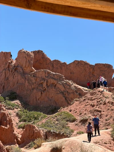 In Tosor, Kyrgyzstan, a landscape of red rock formations rises against a clear blue sky. A group of people stands atop one of the formations, while two individuals walk along a pathway cut into the rock face. Green vegetation clings to the side of the formations. A wooden structure is visible in the foreground.