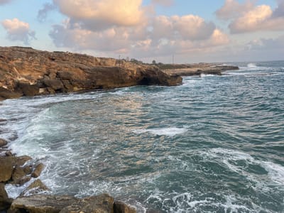 The image depicts a rocky coastline in the Mate Asher Regional Council, Israel, likely near Achziv National Park. The sea is a deep turquoise with whitecaps and foam from the waves crashing against the rocks and the shore. The sky is a pale blue with scattered clouds tinged with the warm hues of sunset. Jagged, brown rocks dominate the left side of the image, forming a rugged cliff face and leading down to the water. In the distance, the coastline continues with more rock formations and a hint of a town or structures further along the shore. The overall scene is one of natural coastal beauty under a dynamic sky.