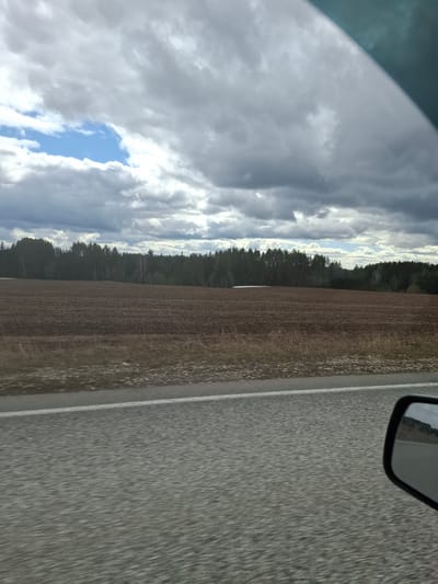 The scene depicts a view from a vehicle, possibly a car, driving on a road. The road appears to be asphalt and has a white line marking the edge. To the left of the road is a brown field. Beyond the field is a line of trees, possibly a forest. The sky is filled with white and grey clouds, with a patch of blue visible. A portion of the vehicle's side mirror is visible in the upper right corner. The scene is rotated to the left, with the road oriented vertically. The location is likely near Ledukhi, Russia, but specific details to verify this claim are not visible.