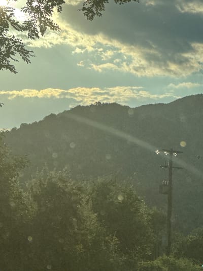 An outdoor, low-angle, medium shot captures a sunlit mountainous landscape under a partly cloudy sky. Trees and foliage frame the bottom and upper left of the scene, with sunlight filtering through leaves at the top. A power pole with multiple lights and a transformer stands near the center right, partially obscured by trees. Sunbeams are visible piercing through the clouds and light haze, illuminating the mountainside. The overall atmosphere is serene and natural, with a slight atmospheric haze. The location is identified as Melivoia, Greece.