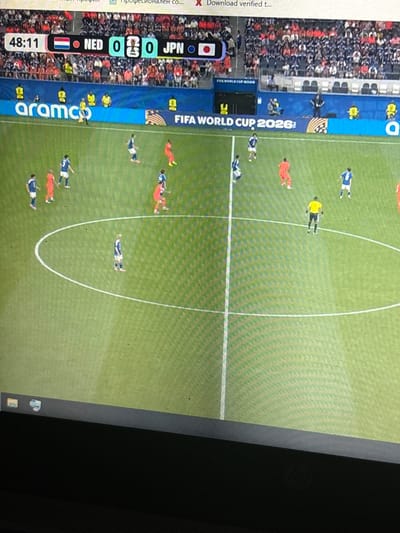 The image is a photograph of a soccer match displayed on a screen, possibly a laptop or television. The screen shows a live broadcast with a score overlay indicating the time is 48:11, with the score being NED 0 - 0 JPN. Flags representing the Netherlands (NED) and Japan (JPN) are visible. A logo for "arama" is displayed on a blue banner in the background, along with "FIFA WORLD CUP 2026". The soccer field is green with white lines. Several players in blue and orange jerseys are visible on the field, along with a referee in a yellow jersey. The image appears to be taken from an angle, with the bottom portion of a dark surface, likely the device the image is displayed on, visible in the foreground. The scene is indoors or in a controlled environment where the screen is being viewed. There is no discernible connection to the city of Enevo, Bulgaria.