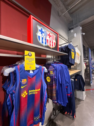 This is a photograph of a retail store interior, likely a sports merchandise shop. The scene depicts shelves filled with blue and red FC Barcelona jerseys and other apparel. A prominent yellow tag hangs from a hanger, reading "CUSTOMIZE IT ON YOUR WAY" and "AVAILABLE AT CASH DESK." In the background, a red-framed display featuring the FC Barcelona crest is visible. Various security cameras are mounted on the wall and ceiling. The flooring is a polished concrete. The environment suggests a commercial setting focused on sports fan apparel. This store is located in La Roca Village, a luxury outlet shopping center in La Roca del Vallès, Spain.